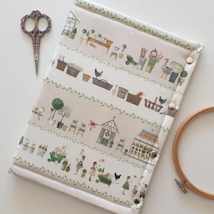 May include: A rectangular fabric needlework project keeper featuring a cream background with a repeating garden-themed print of greenhouses, potted plants, chickens, and gardening tools, secured with three silver metal snaps along the side edge.