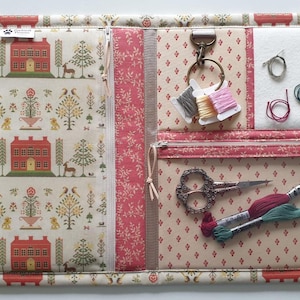Può includere: Una custodia in tessuto per organizzare il cucito con esterno color crema e motivo a case e alberi rossi. L'interno presenta tasche con cerniera, un'area in feltro bianco per aghi, un anello metallico con rocchetti di filo e forbici.