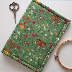 May include: A rectangular padded fabric sleeve in sage green featuring a repeating pattern of houses, bees, flowers, and silhouettes. The sleeve is secured with three silver metal snaps along the side edge. Vintage-style scissors and a wooden embroidery hoop are positioned nearby.