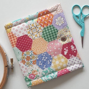Puede incluir: Un estuche de patchwork hecho a mano con parches hexagonales multicolores. El estuche tiene un cierre de botón a presión plateado. También se ven unas tijeras azul claro y un bastidor de bordado de madera.