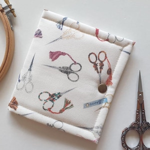 May include: A square fabric case featuring a repeating print of various vintage-style scissors with colorful tassels. The off-white case is secured with a single bronze-colored metal snap button and includes a small blue rectangular label.