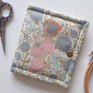 May include: A fabric needle case with a floral and fairy design. The case features a button closure and a quilted border. The illustration depicts a fairy with orange hair and blue wings, surrounded by blue flowers and green foliage.