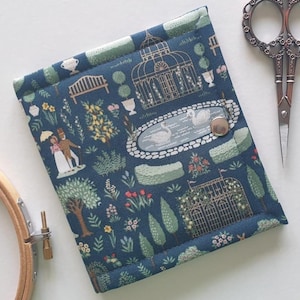 May include: A navy blue fabric needlework case with a garden scene print, featuring a pond with swans, a couple, and a greenhouse. The case is closed with a silver snap. A pair of decorative scissors and an embroidery hoop are next to the case.