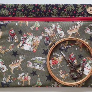 May include: A rectangular fabric pouch with a red zip and a dark green base featuring a festive print of woodland animals, snowmen, and stars. The top border is black with a red star pattern. A wooden embroidery hoop and metal scissors sit on top.