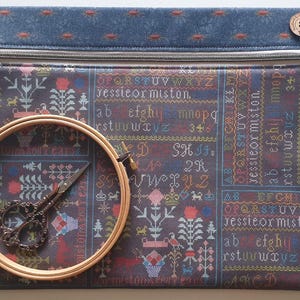 Peut inclure: Une pochette zippée bleu foncé avec un motif de point de croix, un cercle à broder en bois et des ciseaux ornés. La pochette présente des motifs de texte et floraux de différentes couleurs. Un bouton en bois est sur le coin supérieur droit.