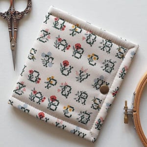 May include: A square fabric case featuring a cream background with a repeating cross-stitch alphabet pattern in dark green, accented by small red, yellow, and blue floral motifs. The case includes a single metallic snap closure on the front.