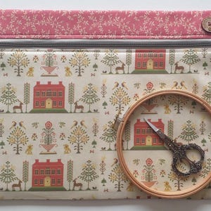 Puede incluir: Una bolsa rectangular de tela con cierre de cremallera, con un estampado crema de casas rojas, árboles y ciervos. Un bastidor de bordado de madera y unas tijeras decorativas descansan sobre la bolsa. La parte superior es un estampado floral rosa.