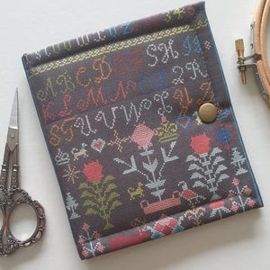 May include: A gray cross-stitch needlework case with a floral and alphabet design. The case has a brass snap closure and is next to a pair of ornate scissors and an embroidery hoop.