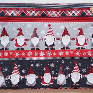 May include: A rectangular fabric pouch featuring a festive gnome design. The pouch has a red zipper and a button closure. The gnomes are depicted with white beards and red hats, holding hands against a gray and black background with snowflakes. The top and bottom borders have a plaid pattern.