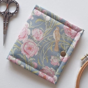 Può includere: Un astuccio per cucito rettangolare e imbottito con un motivo floreale di peonie rosa e un uccello marrone su sfondo grigio. L'astuccio ha una chiusura a bottone e un bordo rosa. Sono visibili un paio di forbici ornate e un telaio da ricamo in legno.