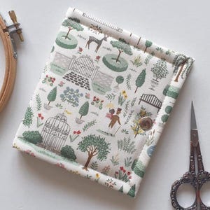 May include: A cream-coloured fabric needle case featuring a garden scene print. The case has a repeating pattern of trees, deer, and people. A silver snap closure is visible. An embroidery hoop and decorative scissors are also in the image.