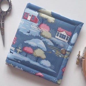 May include: A square fabric case with a blue background featuring a printed landscape of houses, trees, and a white fence. The case has a silver metal snap closure and is stitched along the edges. It is positioned near metal scissors and a wooden hoop.