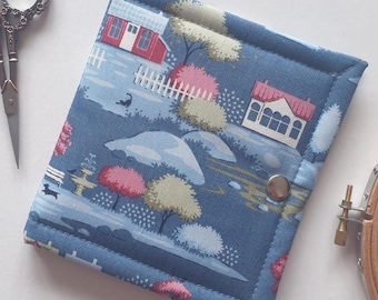 Puede incluir: Un estuche de tela cuadrado con fondo azul que presenta un estampado de casas, árboles y una valla blanca. El estuche tiene un cierre de broche de metal plateado y está cosido por los bordes. Se sitúa junto a unas tijeras y un bastidor de madera.
