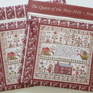 May include: Two rectangular fabric pouches with a floral and striped pattern in burgundy and cream. Each pouch features a cross-stitch design with a house, trees, and text. The top of one pouch reads "The Queen of the May 2026 ~ Ann Watson".