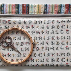 May include: A rectangular fabric zip pouch featuring a cream background with a repeating black cross-stitch alphabet pattern and small floral accents. The top border has a colourful striped floral print. A wooden embroidery hoop and metal scissors.