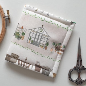May include: A cream-colored, quilted needlework case with a garden-themed print featuring a greenhouse, potted plants, and gardening tools. The case is closed with a silver snap. A pair of ornate scissors and a wooden embroidery hoop are also visible.