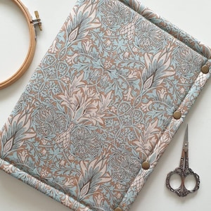 Puede incluir: Un organizador de manualidades rectangular y acolchado con un estampado floral en azul claro, beige y crema. El organizador tiene cierres de botón a presión de color latón. También se ven un bastidor de bordado de madera y unas tijeras decorativas.