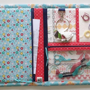 May include: A craft organiser made of fabric with a floral pattern in blue, red, and yellow. It has pockets, a zip, and a ring with embroidery floss and scissors. Designed for cross-stitch and embroidery supplies.
