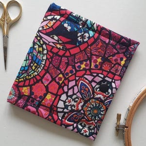 May include: A colorful, patterned fabric book cover with a black snap closure. The cover features a stained-glass-like design in shades of red, pink, blue, and yellow. Gold scissors and a wooden embroidery hoop are visible.