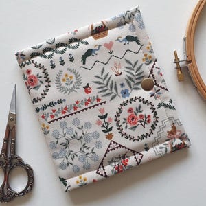 May include: A rectangular fabric pouch featuring a cream background with a multi-coloured botanical and folk-art print including flowers, birds, and hearts. The pouch is secured with a single bronze-coloured metal snap button closure.