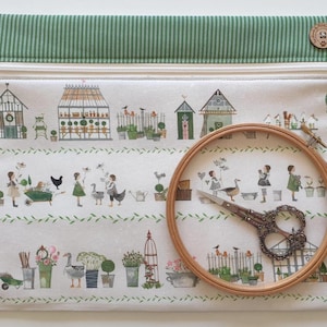 Può includere: Una pochette con cerniera color crema con la parte superiore a righe verdi e un bottone in legno. La pochette presenta una scena di giardino stravagante con illustrazioni di serre, persone e piante. Un telaio da ricamo in legno e forbici decorative sono in primo piano.