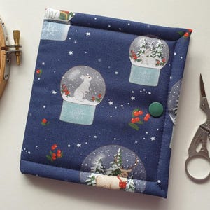 May include: A rectangular fabric case in dark blue featuring a repeating pattern of snow globes containing white rabbits and deer, accented with red berries and white stars. The case is secured with a single green snap button closure.