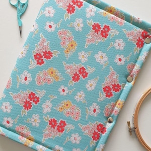 Puede incluir: Un soporte rectangular para proyectos de costura con un estampado floral sobre un fondo azul claro. El diseño presenta flores rojas, rosas y amarillas. Tiene un borde acolchado y cierres de presión metálicos. También se ven unas tijeras turquesas y un bastidor de bordado.