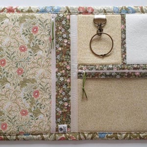 Pode incluir: Um organizador de tecido retangular com um padr&atilde;o floral de flores cor-de-rosa e folhagem verde. Inclui um fecho vertical, um mosquet&atilde;o de metal com anel, um remendo de feltro branco e um bolso horizontal com fecho nas bordas.
