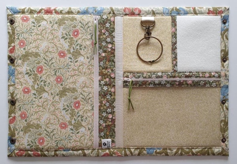 Pode incluir: Um organizador de tecido retangular com um padr&atilde;o floral de flores cor-de-rosa e folhagem verde. Inclui um fecho vertical, um mosquet&atilde;o de metal com anel, um remendo de feltro branco e um bolso horizontal com fecho nas bordas.