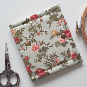 May include: A floral fabric needlework case with a button fastening. The case has a pale green background with a pattern of red, pink, and yellow roses and green leaves. A pair of ornate scissors and an embroidery hoop are also visible.