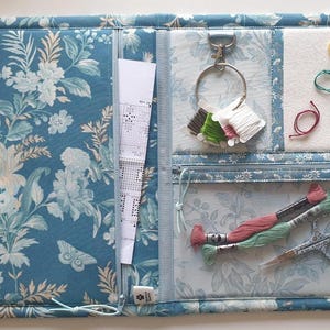 May include: A floral-patterned craft organizer in shades of blue and cream, featuring multiple compartments. It holds embroidery floss, scissors, and a pattern. The organizer has a zippered pocket and a key ring for supplies, ideal for cross-stitch or embroidery.