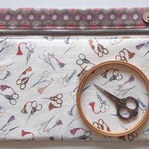 May include: A cream-colored zippered pouch with a pattern of scissors and tassels. A wooden embroidery hoop holds a pair of decorative scissors. The top of the pouch has a patterned border and a wooden button.