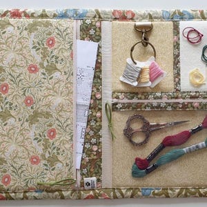 Pode incluir: Um organizador de artesanato em tecido com estampa floral de flores rosas e folhagem verde. Inclui um bolso com z&iacute;per, um anel de metal com tr&ecirc;s carret&eacute;is de linha, uma se&ccedil;&atilde;o de feltro branco, tesoura e duas meadas de linha de bordar.