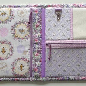 Può includere: Un organizer in tessuto con un pannello bianco a motivi floreali e di api, un dorso floreale color lavanda e un pannello color lavanda con motivo a griglia, dotato di tasca con zip, chiusura girevole in metallo e una sezione in feltro crema.