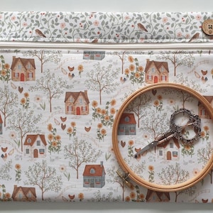 May include: A rectangular fabric pouch with a white zip closure, featuring a pattern of houses, sunflowers and chickens. A wooden embroidery hoop and ornate metal scissors are placed on the pouch. A small wooden button is attached to the top right.