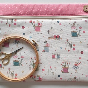 Könnte beinhalten: Eine rechteckige Nähprojekt-Tasche mit einem rosa gepunkteten Oberteil und einem Reißverschluss. Die Tasche zeigt einen skurrilen Druck von Nähmaschinen, Garnrollen und floralen Elementen. Ein hölzerner Stickrahmen mit goldener Schere liegt davor.