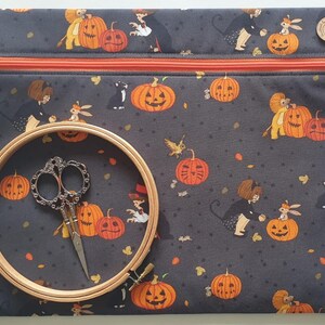 May include: A rectangular dark grey zipped pouch featuring a repeating Halloween pattern of jack-o'-lanterns, children in costumes, and black cats. A round wooden embroidery hoop and ornate silver-toned metal scissors rest on the pouch.