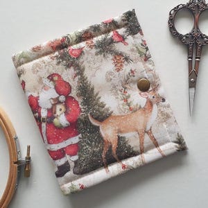 May include: A Christmas-themed needlework case featuring a festive Santa Claus and a deer in a winter scene. The case is beige with a button closure and is next to a pair of decorative scissors and a wooden embroidery hoop.