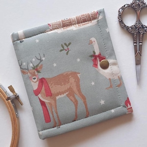 May include: A square fabric needle case in sage green featuring a print of a brown deer wearing a red scarf, a white goose with a red bow, and small white stars. The case is secured with a single brass-colored metal snap button closure.