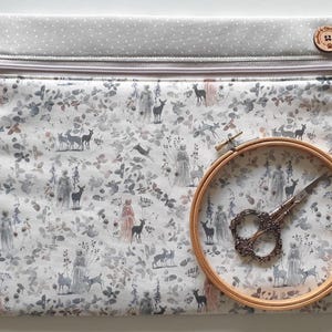 May include: A rectangular fabric pouch with a zipper closure, featuring a woodland pattern with deer and figures in muted tones. A wooden embroidery hoop holds decorative scissors. A wooden button is on the top right.