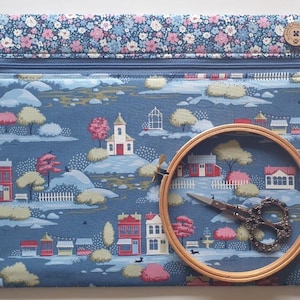 May include: A blue fabric zipper pouch featuring a repeating pattern of houses, trees, and market stalls. The top border has a floral print, and a round wooden embroidery hoop with ornate metal scissors is placed on the front of the pouch.