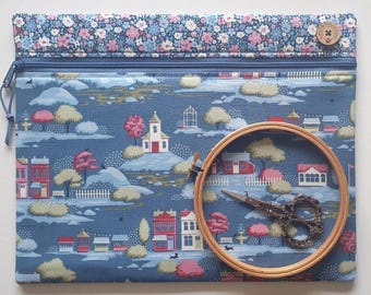 Puede incluir: Un estuche de tela azul con cremallera y un patrón repetitivo de casas, árboles y puestos de mercado. El borde superior tiene un estampado floral, y un bastidor de madera con tijeras de metal ornamentadas se coloca sobre el estuche.