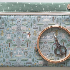 May include: A rectangular fabric pouch with a zipper closure, featuring a light blue garden print with trees, fountains, and figures. A wooden embroidery hoop and ornate metal scissors are placed on top. A green band with rabbit motifs is at the top.
