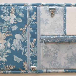 May include: A blue floral fabric organiser with multiple compartments. Features a zip closure, a metal clasp, and a clear pocket. The fabric has a light blue background with white and cream-coloured floral patterns. Metal poppers secure the edges.