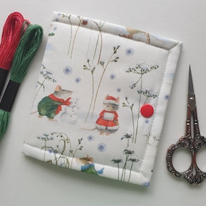 May include: A white, padded needlework case with a whimsical winter scene featuring mice building a snowman. The case has a red button closure and is accompanied by red and green embroidery threads and small scissors.