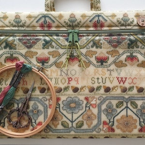 May include: A beige cross-stitch project bag with a green zip and handle. The bag features a detailed floral and geometric pattern. A wooden embroidery hoop holds thread and small scissors.