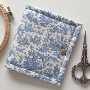Può includere: Un astuccio per cucito in tessuto toile de Jouy blu e bianco con chiusura a scatto in ottone. L'astuccio è accanto a un paio di forbici ornate e a un telaio da ricamo in legno. Il tessuto presenta una scena pastorale.