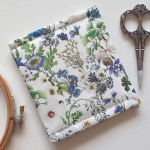 Può includere: Una custodia in tessuto quadrata con sfondo bianco e una stampa botanica dettagliata di fiori selvatici e fogliame blu, bianchi e verdi. La custodia è chiusa da un bottone a pressione in metallo color argento al centro.