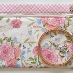 May include: A rectangular fabric zip pouch featuring a white background with a floral pattern of pink roses and blue flowers. The top border is pink and white gingham fabric with a wooden button. A wooden embroidery hoop and scissors are included.