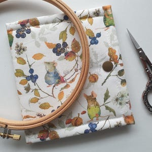 Op de afbeelding: Een witte stoffen borduurorganizer met een grillige print van muizen, bessen en bladeren. Een houten borduurring en een kleine schaar liggen naast de organizer. De organizer heeft een drukknoopsluiting.